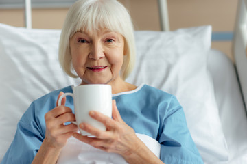 smiling senior woman lying in bed, looking at camera and drinking tea in hospital