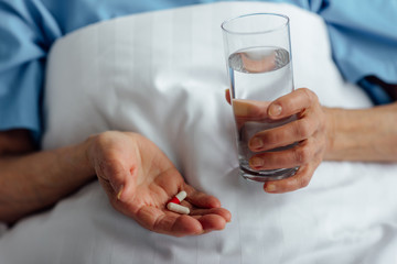 selective focus of senior woman lying in bed and holding pills with glass of water in hospital