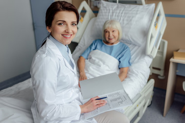 Obraz premium smiling female doctor sitting on bed with diagnosis and looking at camera while senior woman lying in bed in hospital