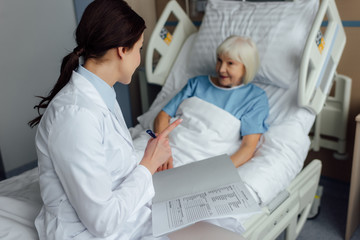 female doctor sitting on bed, holding diagnosis and pointing with finger while senior woman lying in bed in hospital