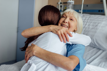Obraz na płótnie Canvas happy senior woman lying in bed and hugging female doctor in hospital