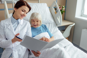 Fototapeta premium beautiful smiling female doctor holding diagnosis and consulting senior woman lying in hospital bed