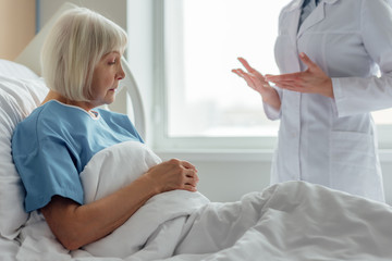 female doctor consulting sad senior woman with grey hair lying in hospital bed