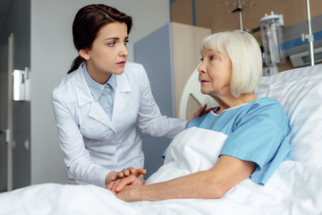 worried female doctor holding hands and consulting senior woman lying in hospital bed