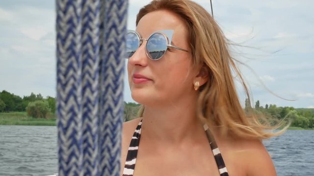 Pretty beautiful girl with light brown hair and wearing sunglasses lies on deck of white luxury yacht in swimsuit and sunbathing at sunday in summer traveling by sea during voyage or cruise.