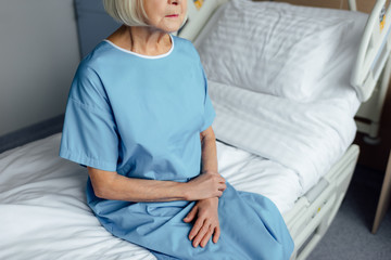 Fototapeta na wymiar cropped view of senior woman with folded hands sitting on bed in hospital
