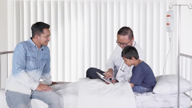 Young male doctor explaining medical record on sick little boy patient while sitting on the bed in hospital room. Shot in 4k resolution