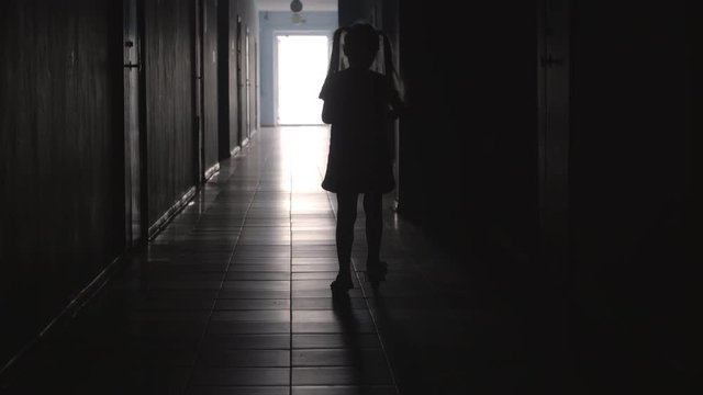 Rear view of silhouette of little girl wearing dress riding a scooter along dark hallway 