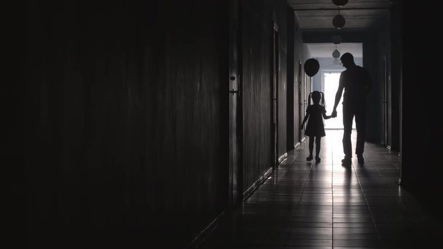 Silhouettes of little girl holding hands with father and balloon while walking towards the camera along hallway