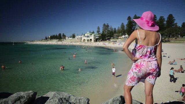 Woman in hat looking at the turquoise waters of Cottesloe Beach, Perth's most famous town beach, Western Australia. Girl sunbathing on the rocks of the Indian Ocean. Summer season with blue sky.