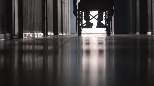 Low section of silhouette of paraplegic man on wheelchair riding away from the camera along hospital hallway