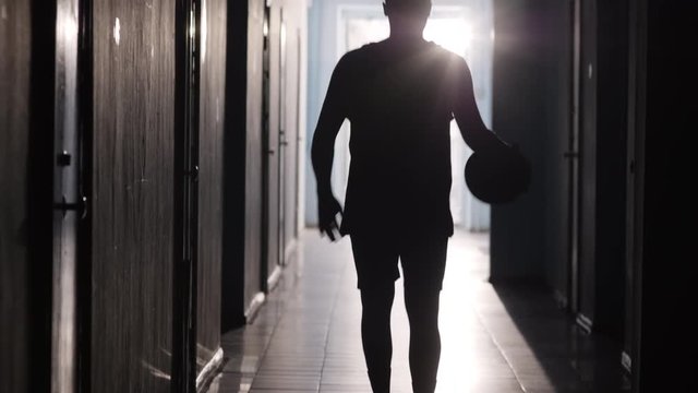 Tilt up shot of silhouette of basketball player in sportswear walking along hallway and dribbling the ball 