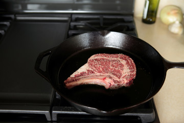 Rib eye steak cooking in a cast iron pan on a natural gas stove top.
