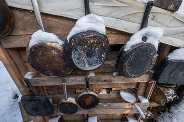 Old pans in burnt ash hanging on wall. A warm looking cottage at the middle of a snowy winter forest. Firewood stacked in piles, covered with snow. cutted trunks in a alpine hut in winter forest