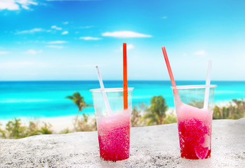 Two strawberry red drift-ice with straw on the beach. In the background is blue sky, palms, sea nd sandy beach. This is situated in tropical resort in Caribbean.