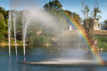 Little rainbow from the water fountain lake, city park pond, water jet beats up against the background of green trees, summer weekend rest