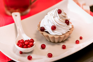 a small cream cake with berries and lingonberry stands on a white plate next to a spoon full of red juicy fresh berries, dessert