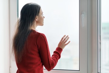 Young woman in red sweater looking out the window, female put her hands on the window glass, side view, window in skyscraper in morning on cloudy day