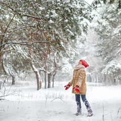 Beautiful young girl wearing a beige down jacket and red knitted hat and scarf playing in a snowy winter park on Christmas day. Child playing with snow in winter. Kid play and jump in snowy forest	