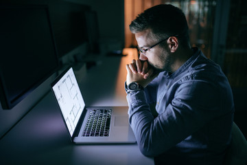 Architect holding head on hands with crossed fingers while working on project late at night. Office interior.