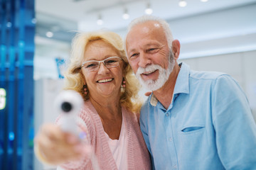 Happy senior couple looking at each other and trying out 360 degrees camera. Tech store interior. Selective focus on couple.