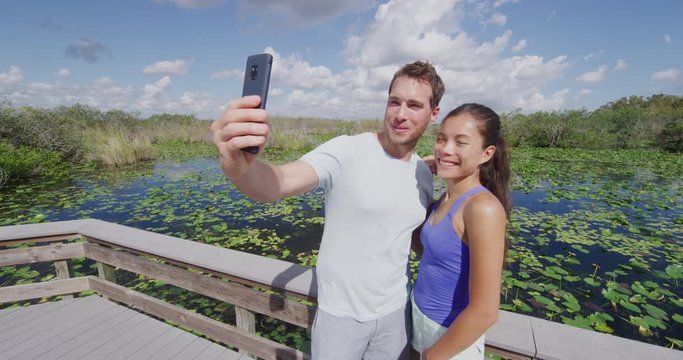 Everglades National Park - tourist couple taking phone selfie photo visiting Florida Everglades. Couple on travel vacation walking Anhinga Trail. Shot on RED in SLOW MOTION.