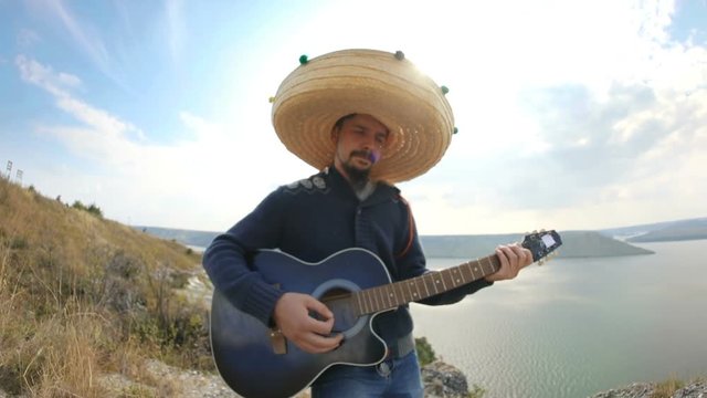 Mexican man wearing sombrero standing on the edge of the hills over the lake and playing acoustic guitar. Funny music performance on sunny day. Fish-eye, close-up.