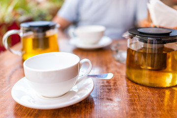 Closeup of two white cups on plates and green or oolong tea in breakfast brunch outdoor cafe restaurant outside wooden table with sunny teapot or pot