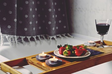 Glass with alcoholic drink, strawberry and candles for romantic date on bath caddy. Romantic bath for Valentine's Day