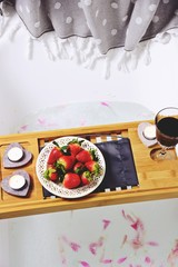 Glass with alcoholic drink, strawberry and candles for romantic date on bath caddy. Romantic bath for Valentine's Day