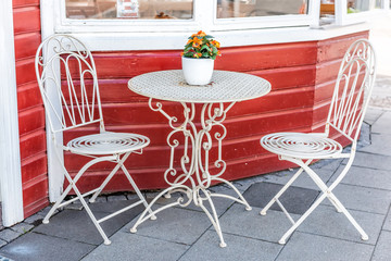 White empty metal table outside restaurant cafe with two chairs on sidewalk street and orange flowers in flowerpot potted plant setting with nobody by colorful red building