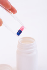 Plastic tweezers in hand putting two-colored pill into open plastic bottle on white background.