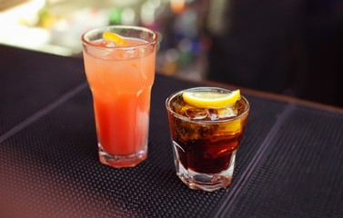 Tequila sunrise and Cuba libre classic cocktails on bar. Close-up cold cocktail with crushed ice and slice of lemon on glass.