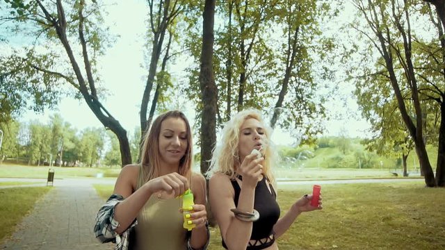 Cheerful hipster girls with sunglasses having fun making bubbles in park outdoors in slow motion