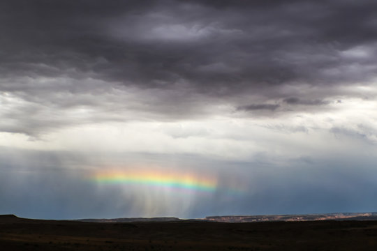 Horizontal rainbow across the middle of a rain squall near the horizon in desert with distant cliffs lit up and foreground dark - very dramatic stormy sky near Salt Lake USA - Powered by Adobe