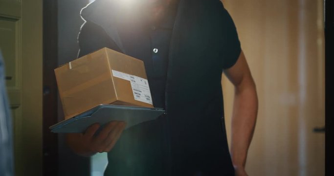 Happy young woman receiving a parcel from mailman delivered in her home. Shot with RED camera i 8K. Concept of courier, delivery, e-commerce, online shopping