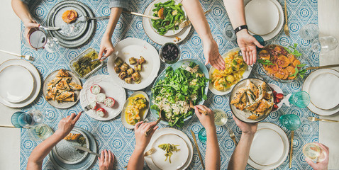 Mediterranean style dinner. Flat-lay of table with salads, starters, pastries over blue table cloth with hands holding drinks, sharing food, top view. Holiday gathering, vegetarian party concept