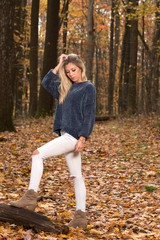 Adorable young blonde woman in blue sweater and jeans stands in autumn woods looking down in thought