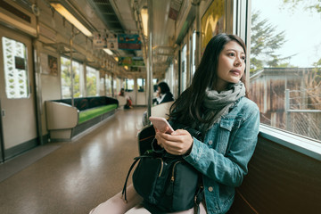 asian travel woman inside subway train looking out beautiful view from window. young girl holding smart phone sitting on seat in metro. fashion chinese woman commute in tour trip wear scarf spring