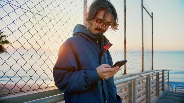 Young millennial hipster man skater and extremal with red glasses using mobile phone or smartphone application or app checking social network news feed to be up to date and being connected to internet