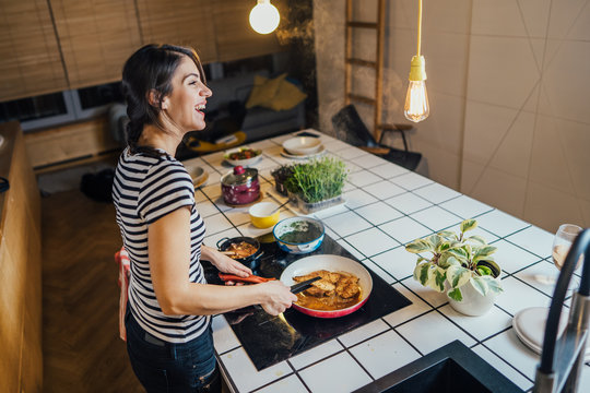 Young woman tasting a healthy meal in home kitchen.Making dinner on kitchen island standing by induction hob.Preparing fresh vegetables,enjoying spice aromas.Eating in.Passion for cooking.Keto diet