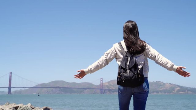 Happy woman backpacker with open hands against blue sea and sky background. Person having fun on summer vacation freedom and imagination concept. carefree girl traveler sightseeing golden gate bridge