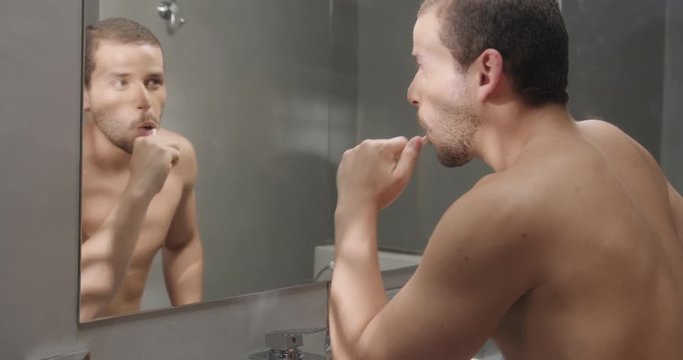 Young hispanic people, male beauty. Latino person in bathroom at home for morning routine, body care. Caucasian gay man brushing teeth, looking at mirror in hotel room on vacation travel. Slow motion