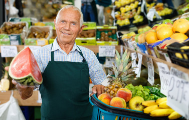 Positive senior male owner of greengrocery shop in apron offering fresh fruits and vegetables