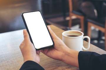 Mockup image of hands holding white mobile phone with blank screen with coffee cup on wooden table in cafe