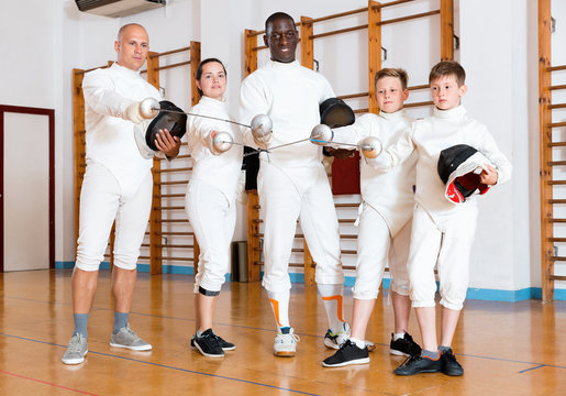 Group portrait of young fencers with coaches holding rapiers in training room