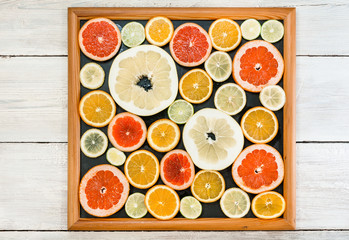 Citrus slices in picture frame. Fresh citrus fruits in different colours and sizes, cut into slices and laid out in a wooden black frame. White wooden background.