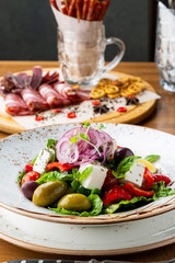 Greek salad with feta cheese, olives and herbs, served in a white plate on a table in a restaurant.