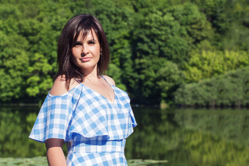 Young woman with dark hair stands against the background of a forest and lake. Sunny summer day