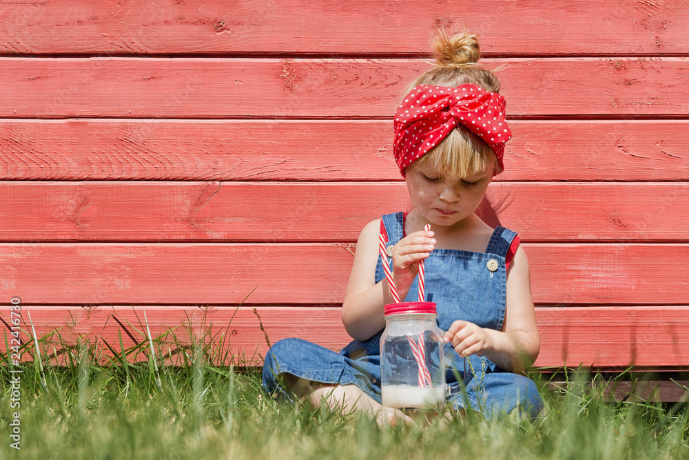 Wall mural little girl in blue dungarees sitting and drinking milk in the garden.